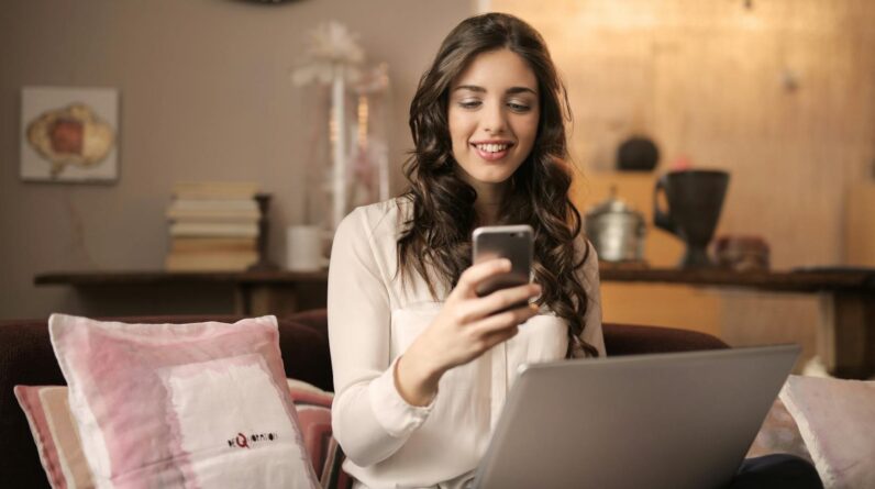 Woman Sitting on Sofa While Looking at Phone With Laptop on Lap